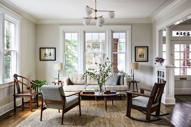 A bright, refined living room with tall multi-pane windows, pale walls, detailed crown moulding and traditional trim. The room is furnished with a light sofa, mid-century-inspired timber armchairs, a low oval coffee table, table lamps, framed artwork and a woven rug. A classic white fireplace surround and built-in shelving are visible on the right, with a glimpse into an adjacent space.