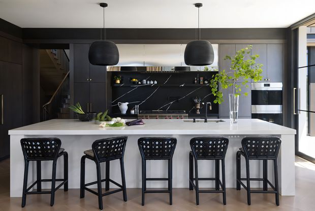 A refined contemporary kitchen dominated by a large white island with seating for five. Dark floor-to-ceiling cabinetry frames a black marble-look splashback and open display shelving, while two oversized black pendant lights hang above the island. Integrated appliances, a dark sink and tapware, timber flooring, and a bright glazed opening complete the space.