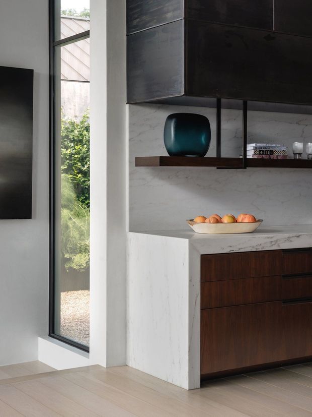Partial view of a refined modern kitchen with dark timber lower cabinetry, a pale veined stone benchtop and matching full-height splashback, and dark metal overhead cabinetry. A slim black-framed vertical window brings in garden light beside the kitchen, while an open timber shelf displays a sculptural dark vase and books. The space has a restrained, high-contrast palette and clean architectural lines.
