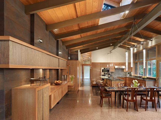 Warm open-plan kitchen and dining interior with extensive timber cabinetry and wall panelling, a long dining table, polished brown-toned floor, and a dramatic exposed-beam timber ceiling. The kitchen includes a central island, integrated appliances, pendant lighting, and large glazed windows. The space has a refined mid-century-inspired character with strong natural timber detailing.