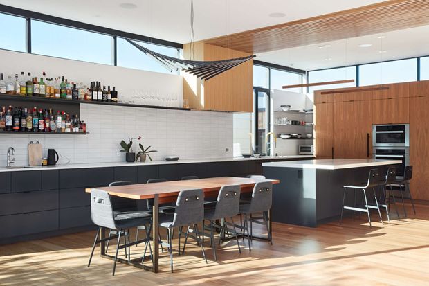 A bright, open-plan contemporary kitchen and dining area with a long island, integrated appliances, extensive black lower cabinetry, and warm timber cabinetry and ceiling detailing. A large dining table with upholstered chairs sits in the foreground, while open shelving displays bottles and glassware along a white tiled backsplash. Expansive windows provide abundant natural light, and a sculptural dark pendant hangs above the dining area.