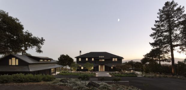 Exterior architectural photograph at dusk showing a dark, low-pitched roofed multi-storey house with extensive rectangular windows, a central entrance, and a separate matching wing to the left. The buildings are set within landscaped grounds and connected by paved driveways and paths, with mature trees framing the composition.