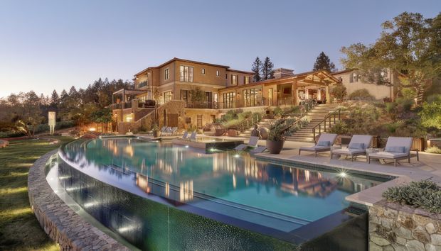 A large luxury multi-level residence viewed from the landscaped pool area at dusk. The property features warm-toned rendered and stone façades, extensive glazing, covered outdoor living areas, broad stairways, terraced landscaping, and a large curved swimming pool with an infinity-style edge. Lounge chairs and built-in outdoor spaces surround the pool.