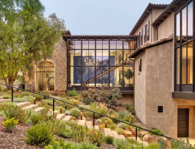 Exterior view of a multi-level residence with warm stucco walls, stone masonry, terracotta-tiled roof edges, and extensive black-framed glazing. A glazed central link reveals an interior stair and chandelier, while landscaped terraced planting and a stepped garden path lead toward the building.