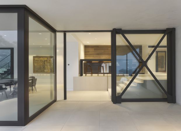 A refined contemporary interior featuring a glazed black-framed partition and large hinged glass door with prominent diagonal bracing. The space has pale large-format flooring, white walls and ceiling, recessed lighting, and warm timber wall cladding in the background. A dining or work area and artwork are visible through the glazing.