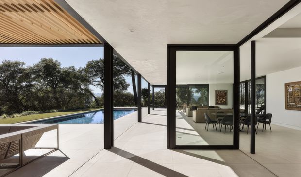 A contemporary indoor-outdoor living area with expansive black-framed glazing opening onto a long swimming pool and landscaped grounds. The covered terrace has pale tiled flooring, a slatted timber ceiling section, and minimal outdoor furniture; through the glazing, a bright dining and living space with black-framed windows and artwork is visible.