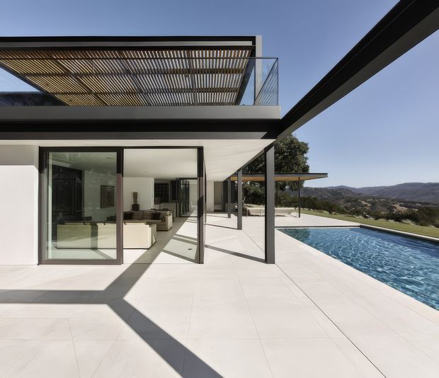 A contemporary two-level house exterior with expansive glass sliding doors, broad cantilevered rooflines, and a rooftop pergola or slatted sunshade. The pale paved terrace extends around a long rectangular swimming pool, with a living area visible through the glazing. The property overlooks a dry, hilly landscape under a clear sky, and strong structural shadows add graphic contrast.