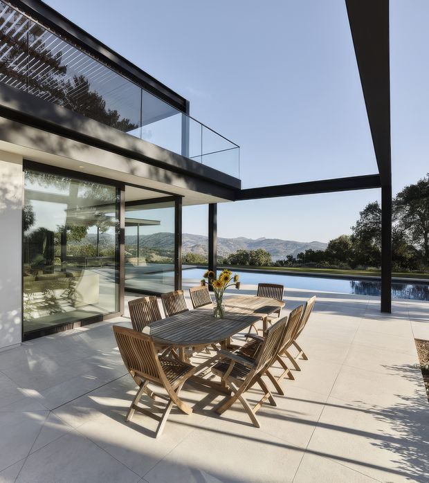 A modern outdoor dining terrace beside a long rectangular swimming pool, framed by a cantilevered two-storey glass-and-dark-metal house. A timber dining table with folding chairs occupies the pale paved terrace, while expansive glazing opens toward rolling hills and mature trees. Clean horizontal rooflines, slender dark structural posts, transparent balustrades, and strong sunlight create a refined indoor-outdoor setting.