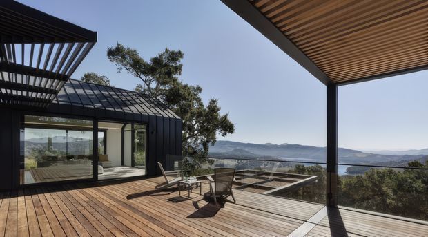 A modern outdoor terrace and deck overlooking a broad mountainous or valley landscape. The space features warm timber decking, a large slatted timber overhead canopy, slim dark metal structural framing, extensive glass walls and balustrades, and outdoor seating. A glazed pavilion or interior living area is visible to the left, while the terrace is the dominant subject.