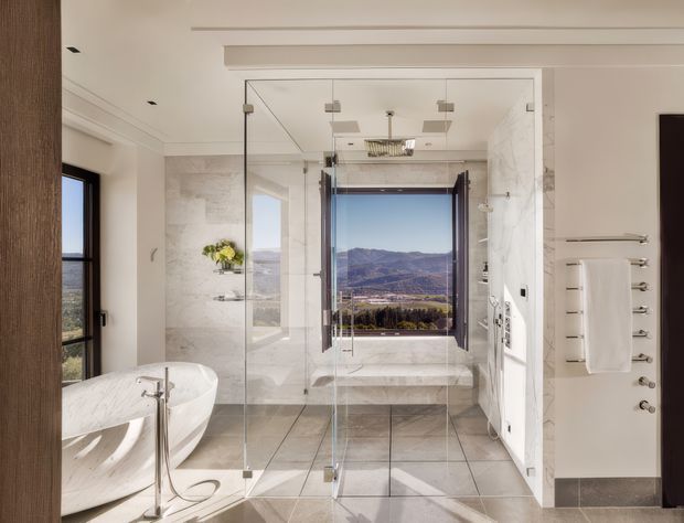 Bright, spacious contemporary bathroom with a freestanding white sculptural bathtub, large frameless glass shower enclosure, pale marble-look wall surfaces, and large grey floor tiles. A wide window offers a distant mountain and valley view, while white built-in cabinetry and towel storage line the right side. The room has abundant natural light, clean lines, and a refined neutral palette.