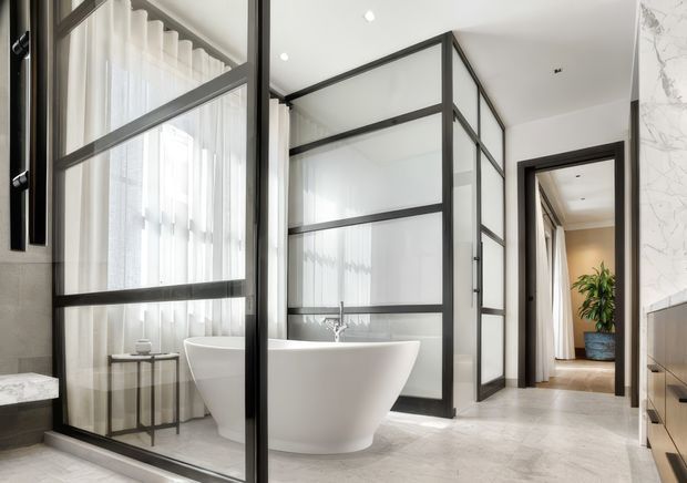 A bright, refined bathroom featuring a freestanding white bathtub enclosed by black-framed translucent glass partitions. Large sheer-curtained windows provide abundant natural light, while pale stone-look surfaces and dark-framed glazing create a contemporary monochromatic scheme. A vanity or cabinetry edge is visible at right, and a doorway leads toward an adjacent area with a potted plant.
