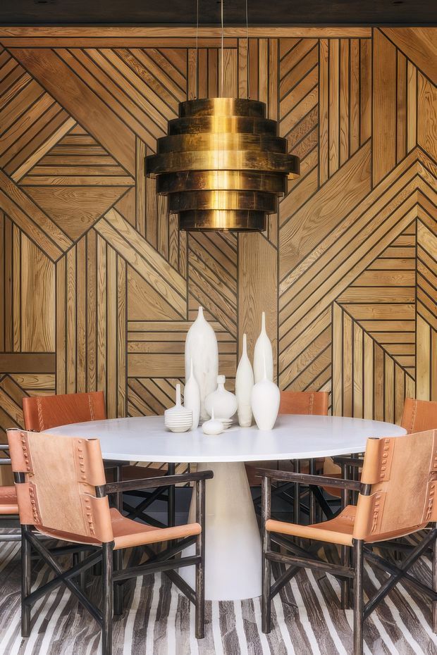 A styled dining area centred on a round white pedestal table with sculptural white vases and several leather dining chairs. The room features a dramatic geometric patterned timber wall with dark outlining, a large tiered brass pendant light, and a striped monochrome rug. The composition is richly layered, warm, and design-focused.