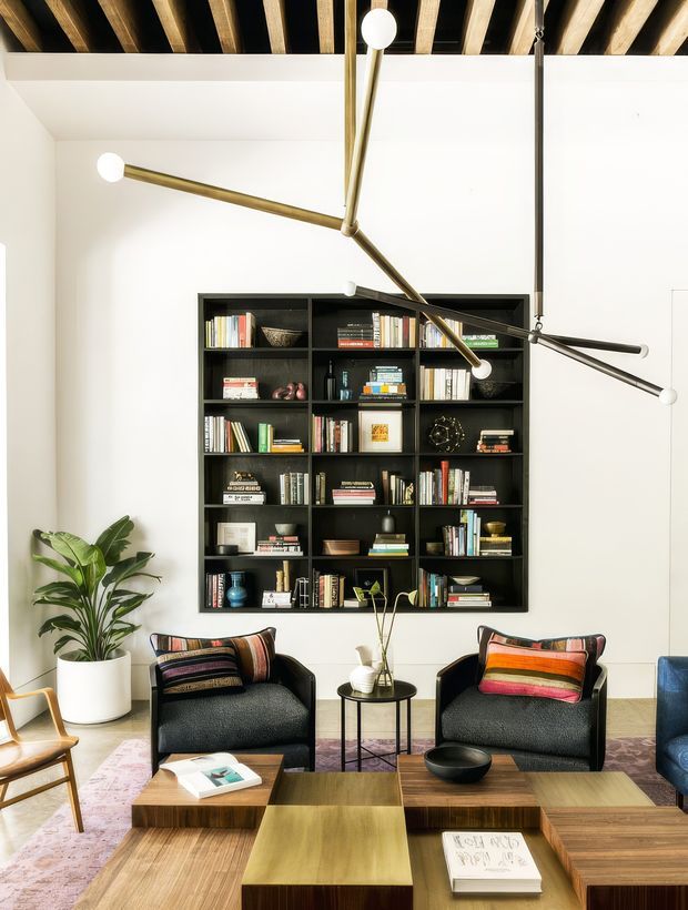 A bright, refined living room with a large black built-in open bookshelf, two dark upholstered lounge chairs, layered wooden coffee tables, and a potted plant. The white walls contrast with a dramatic exposed timber-beam ceiling and sculptural brass-and-black multi-arm pendant lighting. Colourful cushions, books, and restrained decorative objects add warmth and visual interest.