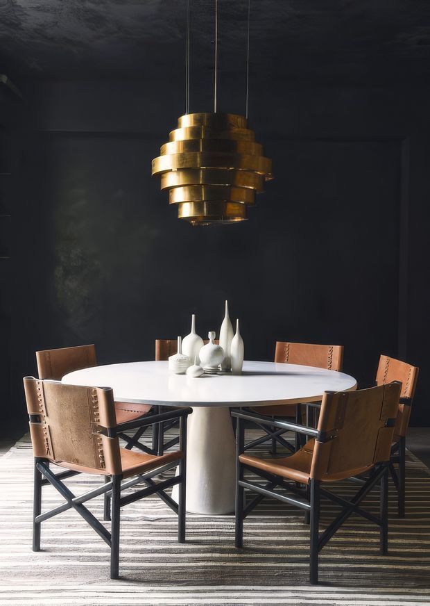 A moody, sophisticated dining room with a round white dining table surrounded by tan leather sling-back chairs with dark cross-braced frames. A sculptural brass pendant light hangs centrally above the table, while white ceramic vases provide a restrained tabletop arrangement. The room has near-black walls and ceiling, dramatic directional lighting, and a large striped area rug.