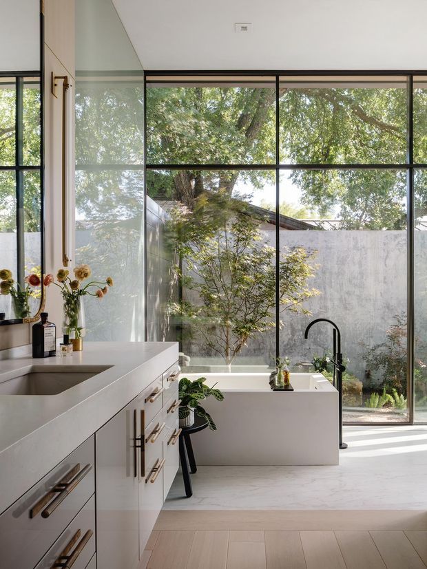 A bright, refined contemporary kitchen with a pale stone island and integrated sink, light cabinetry with long brass-toned handles, and a black gooseneck tap. Full-height black-framed glazing opens the room visually to a planted courtyard, while a raised rectangular planter or built-in bench sits against the window. The space has a clean, warm-minimal palette with abundant natural light.
