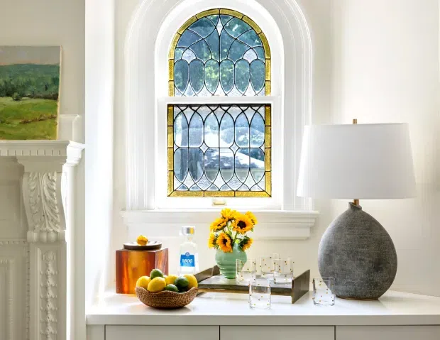 Bright, refined interior vignette centred on a tall decorative stained-glass window with blue, green and yellow geometric detailing. A white carved mantel is visible at left, while a white console or sideboard below the window displays a large textured grey lamp, flowers, fruit and glassware. The space has ornate traditional architectural detailing and a calm, elegant atmosphere.