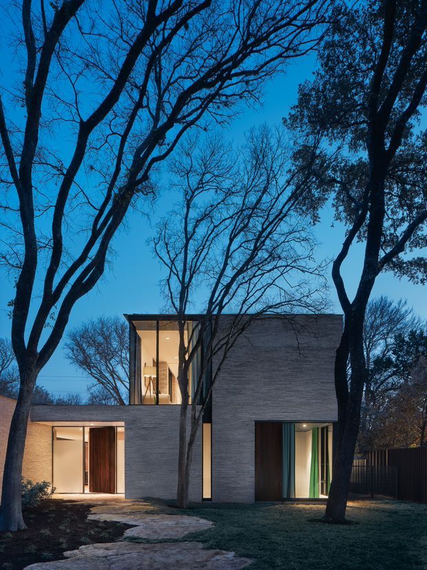 A dusk exterior photograph of a contemporary two-storey residence set among mature trees. The house has a strongly rectilinear composition, dark horizontal masonry or textured cladding, expansive warm-lit glazing, recessed entries, and a projecting upper volume. A stepping-stone path crosses the landscaped front garden toward the entrance, while tall trees frame the building.