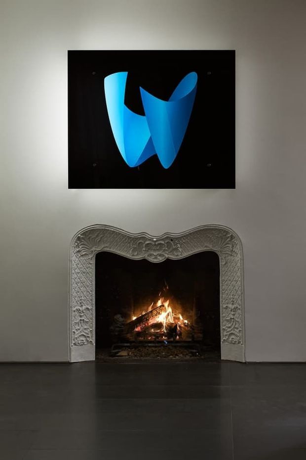 A dim, refined interior focused on an ornate carved fireplace surround with a live fire. Above it hangs a large black artwork featuring a vivid abstract blue ribbon-like form. The walls are painted a soft neutral grey, and the floor is a dark, smooth finish. The composition combines historic decorative detailing with contemporary artwork.