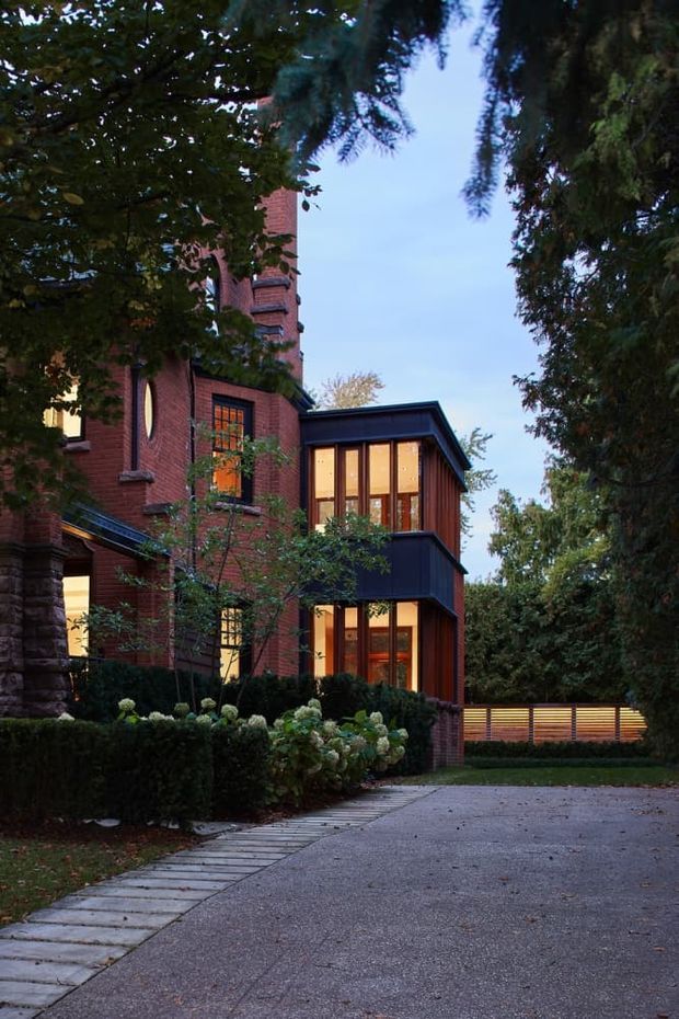 Exterior view of a substantial red-brick residence set within mature landscaping. The image shows a historic-looking brick main house with tall, narrow windows and decorative masonry details, connected to a contemporary dark-clad glazed extension with warm interior lighting. A paved driveway and stone-paver walkway lead toward the house, with hedges, hydrangeas, trees, and a timber privacy fence framing the scene.
