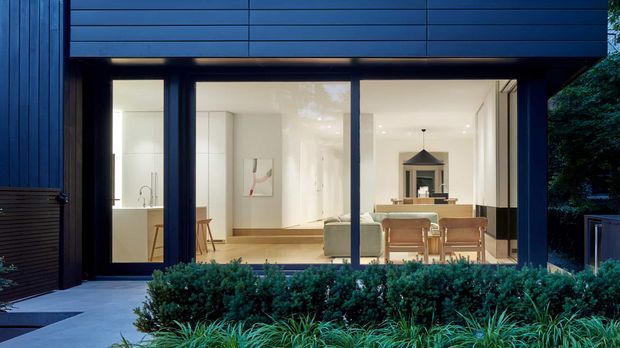 Dusk exterior view of a contemporary, dark-clad single-storey residence. Floor-to-ceiling glazing reveals a bright open-plan interior with a kitchen, dining area, and living space, while dense planted landscaping and a paved entry terrace frame the facade.