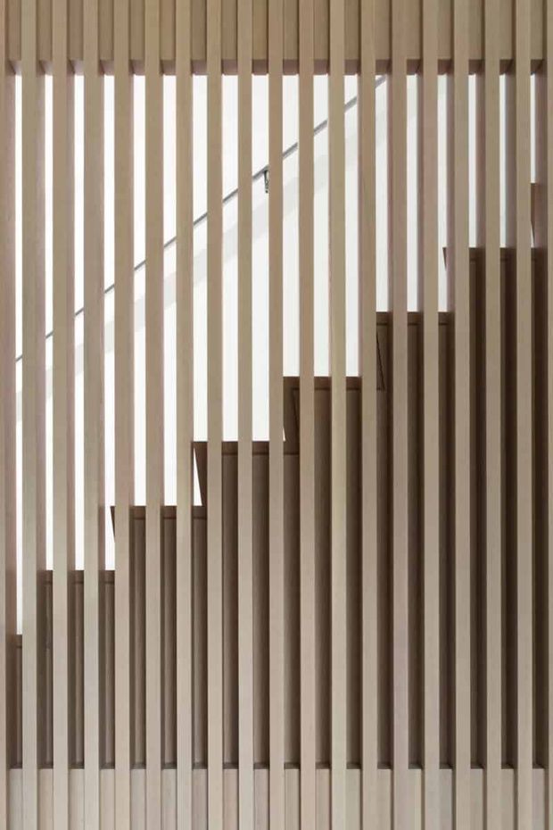 Close-up architectural interior detail of a staircase screened by evenly spaced vertical timber slats. The warm wood battens create a rhythmic geometric partition, with stair treads and a simple handrail visible behind them. The image is minimal, softly lit, and focused on the timber stair enclosure rather than a complete room.