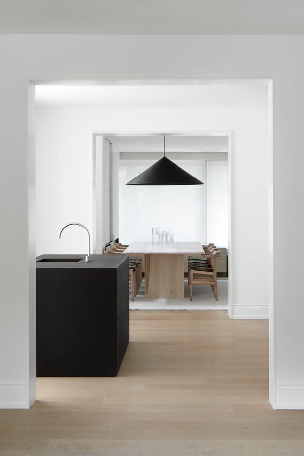 A minimalist open-plan interior viewed through a wide doorway, with a compact black kitchen island and curved stainless-steel tap in the foreground. Beyond it is a light timber dining table surrounded by timber chairs, beneath a large black geometric pendant light. White walls, crisp trim, and pale timber flooring create a restrained, bright composition.