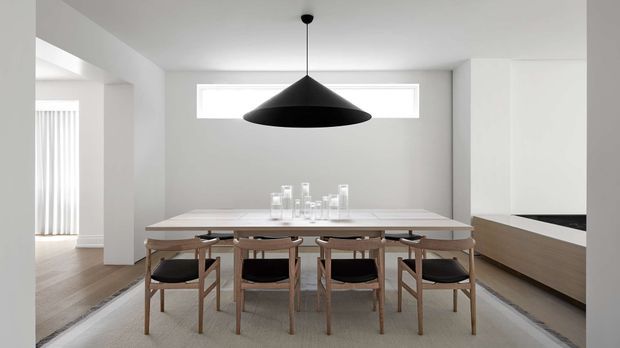 Bright minimalist dining area with a long light-toned dining table and six wooden chairs, positioned beneath a large black cone-shaped pendant light. White walls, a narrow horizontal window, pale timber flooring, and streamlined wood-and-white built-in cabinetry create a clean, restrained interior. A kitchen area is visible to the right and an adjacent bright room is visible to the left.
