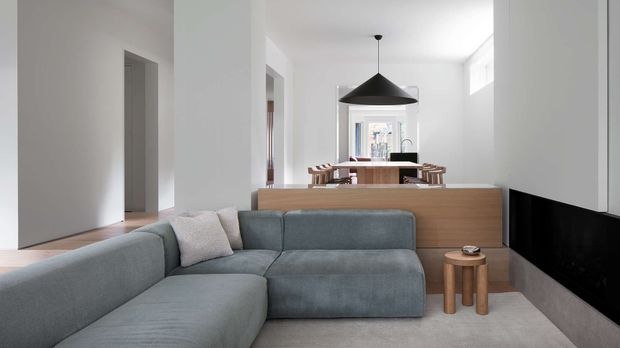 Bright open-plan contemporary living and dining area. A large grey sectional sofa occupies the foreground, facing a sleek black built-in media unit. Behind it is a timber-toned kitchen or dining island with stools, a wooden dining table, and a dramatic black pendant light. White walls, pale flooring, minimal detailing, and abundant natural light create a clean, refined interior.