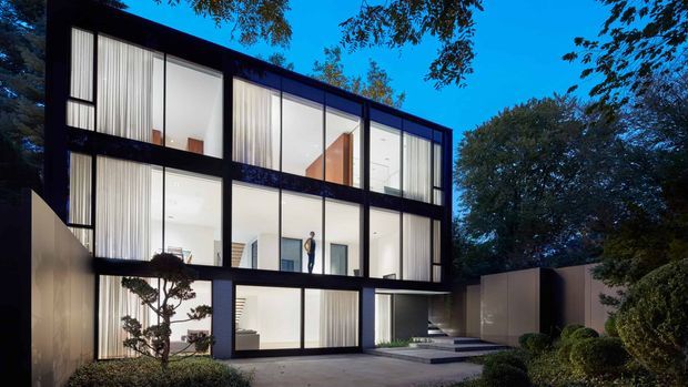 A contemporary multi-storey residence photographed at dusk, with a strongly rectilinear black structural grid and extensive floor-to-ceiling glazing. Sheer curtains, bright minimalist interiors, a recessed entry, paved approach, and carefully landscaped garden surround the house. Mature trees frame the property.