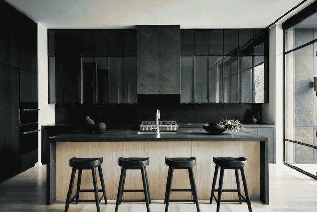 A dramatic contemporary kitchen dominated by matte black cabinetry and dark reflective splashback surfaces. A large island with a light timber-panelled front and dark benchtop is paired with four black bar stools. The kitchen includes a central cooktop and rangehood, integrated appliances, black pendant-free cabinetry, and extensive black-framed glazing on the right that brings in natural light. The restrained palette and strong contrast create a refined, moody atmosphere.