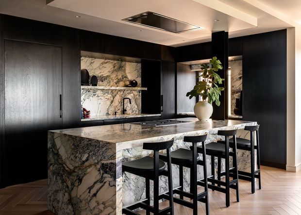 A sophisticated contemporary kitchen dominated by dark, floor-to-ceiling cabinetry and dramatic veined marble surfaces. A large waterfall island with integrated cooktop and seating for four occupies the foreground, while a marble backsplash, open shelving, concealed appliances, black pendant-free stools, and a sculptural ceramic vase with greenery complete the refined, high-contrast composition. The ceiling includes a flush-mounted rangehood and recessed lighting.