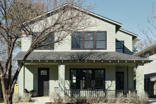 Exterior view of a two-storey residential house with pale horizontal weatherboard cladding, dark-framed windows and doors, a pitched gable roof, and a covered front porch. Mature deciduous trees and low landscaping frame the facade.