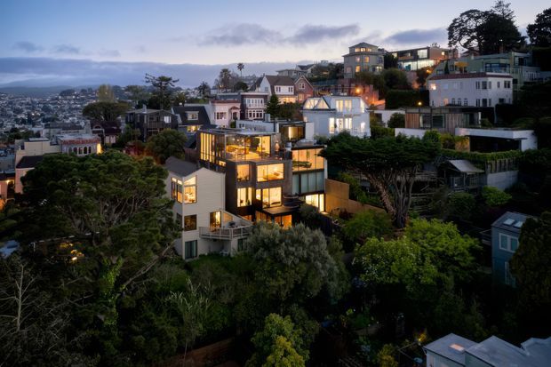 Twilight aerial view of a densely built hillside residential neighbourhood, featuring a prominent contemporary multi-level home with extensive illuminated glazing, balconies, and dark-and-light exterior finishes. Traditional and modern houses step up the landscaped slope behind it, with a broad urban landscape visible in the distance.