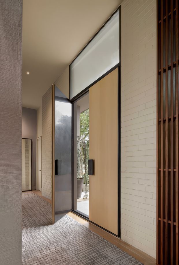 A refined residential entryway or hallway with a large pivot-style timber front door framed in dark metal, translucent upper glazing, and a tall glazed sidelight. Pale brick walls, warm neutral plaster, textured patterned carpeting, and a dark timber vertical-screen feature create a calm, contemporary entrance. A courtyard or garden is visible through the open doorway.