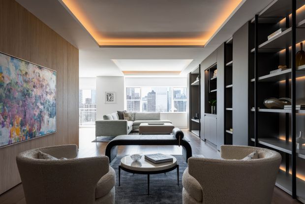 Contemporary living room with a sophisticated, warm-neutral palette. The room features a grey sofa and upholstered lounge chairs arranged around layered coffee tables, a large abstract artwork on a wood-paneled feature wall, dark built-in shelving with integrated warm lighting, and floor-to-ceiling glazing overlooking a dense city skyline. Recessed perimeter ceiling lighting adds an ambient glow.