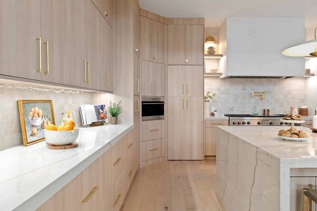Bright, refined contemporary kitchen featuring light timber cabinetry with slender brass pulls, white veined stone benchtops, a pale tiled backsplash, integrated appliances, a freestanding stainless-steel range, and a large island. Warm pendant lighting and carefully styled countertops create an inviting, sophisticated atmosphere.