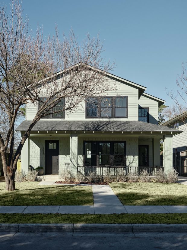 Front exterior view of a two-storey suburban house with pale horizontal weatherboard cladding, dark-framed windows, a gabled upper roof, broad front porch, and a small landscaped lawn with a concrete path leading to the entrance. Leafless trees partially frame the facade.