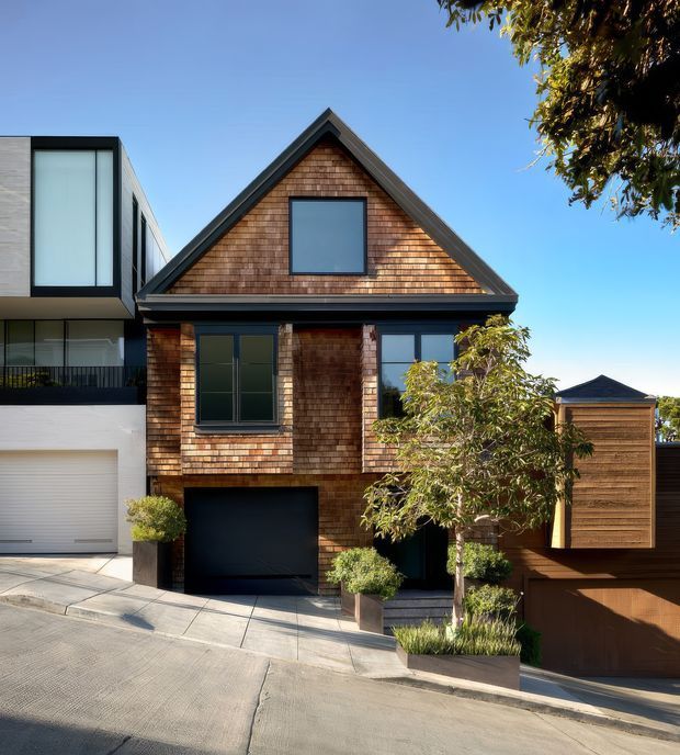 Street-facing exterior of a compact multi-storey townhouse with a steep front-facing gable roof, dark-framed windows, and warm timber shingle cladding. A recessed dark garage sits at ground level, with a modern flat-roofed neighbouring extension on the left and a timber-clad privacy structure on the right. The property is set on a sloping paved street with landscaped planters and trees under clear daylight.