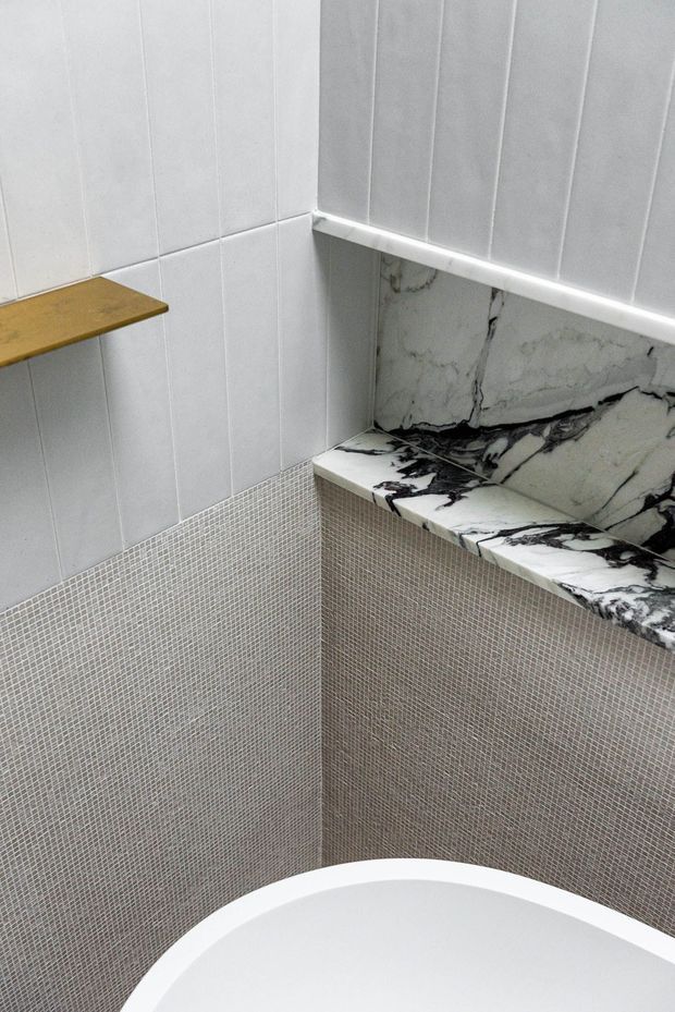 Close-up view of a contemporary bathroom corner featuring a white freestanding bathtub, small mosaic wall tiles, larger white wall tiles, a dramatic black-and-white veined stone ledge and backsplash, and a small timber wall shelf.