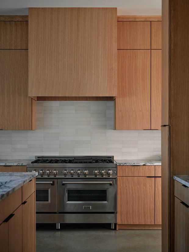 A compact, vertically framed kitchen with warm wood-grain cabinetry, a large concealed range hood enclosure, pale horizontally laid rectangular tile backsplash, grey stone benchtops, and a substantial stainless-steel freestanding range cooker. The space is viewed through a doorway or opening, with additional cabinetry and benchtops in the foreground. The design is restrained and contemporary with natural timber tones and muted grey finishes.
