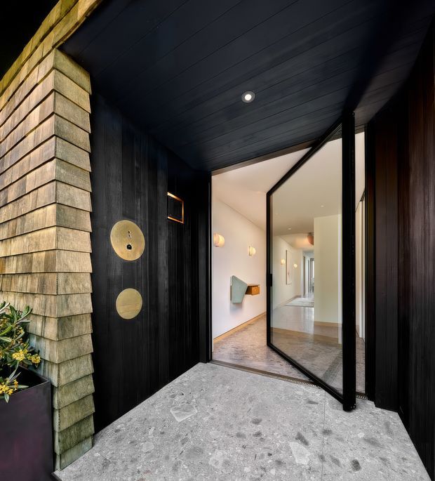 A dramatic covered residential entryway viewed from outside, with a broad pivot-style glazed door opening into a bright interior hall. The entrance combines black vertical timber cladding and a dark timber-lined soffit with a contrasting weathered shingle-clad wall and pale textured concrete paving. Circular brass-toned door hardware and a small planter add detail. The interior beyond has white walls, timber flooring, and understated wall lighting.