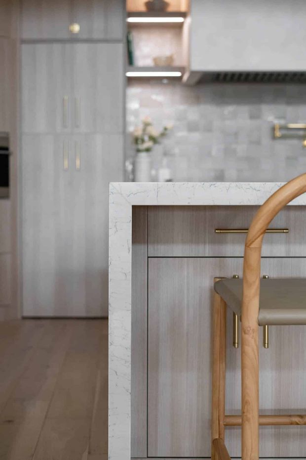 Close-up view of a refined contemporary kitchen featuring tall grey wood-grain cabinetry, a large island with a white veined stone benchtop and integrated grey cabinetry, brass hardware, grey tiled splashback, timber flooring, and a partially visible timber-framed bar stool. The image focuses on the kitchen island and built-in joinery.