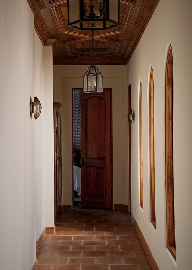A narrow, warmly lit traditional hallway with terracotta tile flooring, cream plaster walls, richly stained timber trim and a coffered timber ceiling. Tall arched timber-framed openings line one side, while a dark carved timber door and lantern-style pendant anchor the far end. Decorative wall sconces and timber cabinetry add historic character.