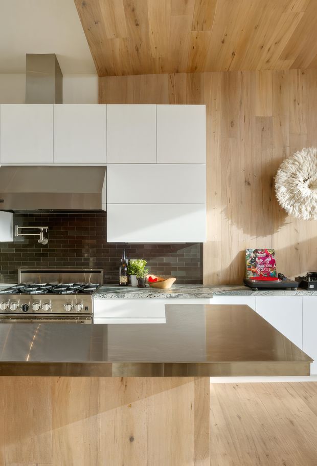 A contemporary kitchen with white flat-panel cabinetry, a large stainless-steel island and range hood, dark glossy rectangular tile backsplash, and extensive natural timber wall and ceiling lining. The island includes a broad work surface and seating overhang, while daylight creates strong shadows across the warm wood surfaces.