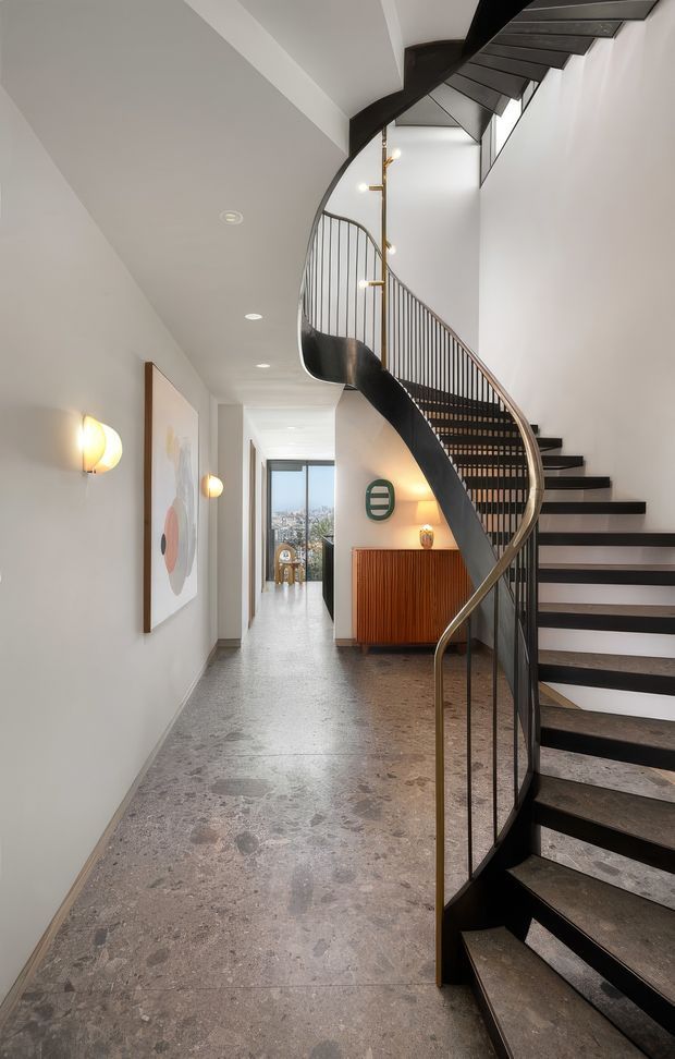 A refined residential hallway dominated by a sculptural, curved staircase with dark treads, black balustrade elements and a slender brass-toned handrail. The corridor has pale walls, recessed ceiling lights, warm wall sconces, a large artwork, a fluted timber console, and a terrazzo or stone-look floor. A glazed opening at the end provides a distant city view.