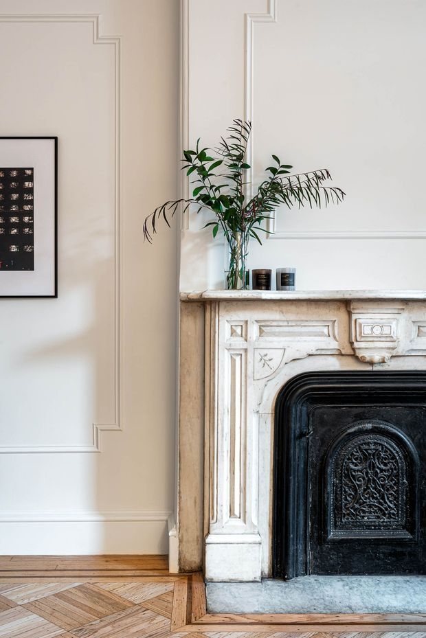 A refined interior detail featuring an ornate carved white fireplace with a black cast-iron fire surround, framed by decorative wall panelling and moulding. A vase of leafy branches and small candles sit on the stone mantel, while patterned parquet timber flooring and framed artwork complete the composition. The image has a bright, elegant heritage character.