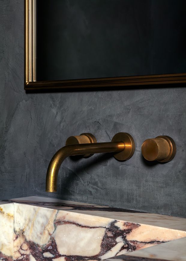 Close-up of a refined bathroom vanity area featuring a wall-mounted aged bronze tap and matching cylindrical control, set against a dark textured plaster wall. A richly veined marble benchtop is visible below, with the lower edge of a dark framed mirror above.