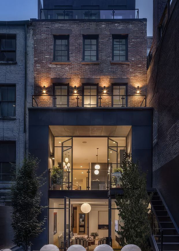 A dusk exterior view of a narrow multi-storey urban building. The facade combines exposed weathered red brick on the upper levels with dark metal cladding around a glazed lower-level extension. Large industrial-style steel-framed windows and doors open onto small balconies with black metal balustrades. Warm interior lighting and wall-mounted exterior lights create a welcoming glow, while rooftop glazing and dense neighboring buildings emphasize the compact city setting.