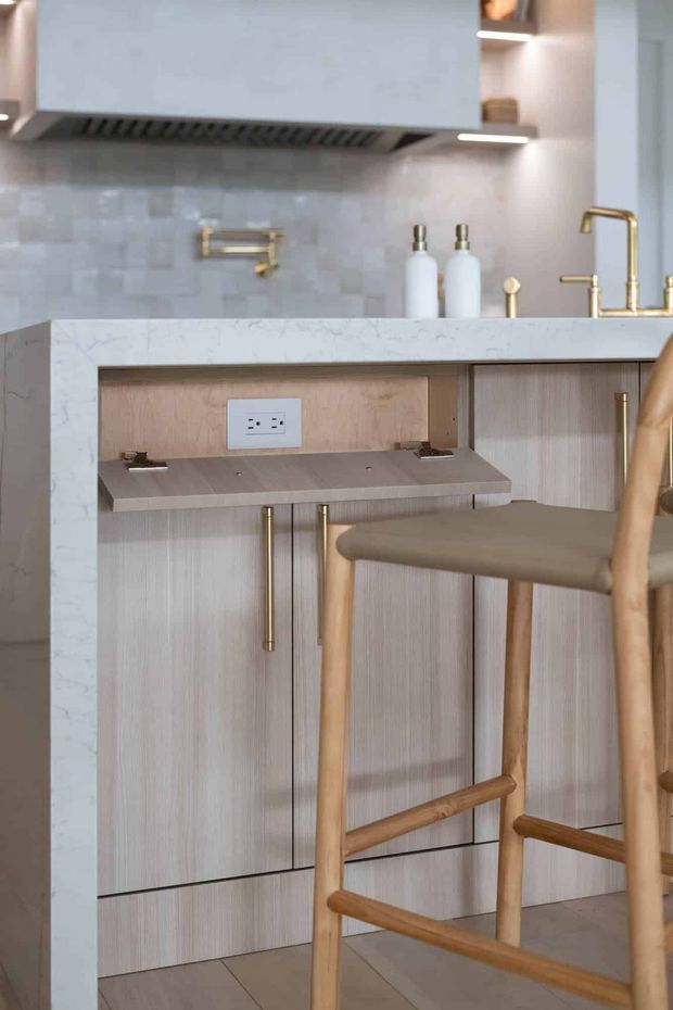 Close-up view of a refined contemporary kitchen, focused on a marble-look island or peninsula with light timber cabinetry, a recessed work surface and power outlet, brass hardware and tapware, pale tiled splashback, and warm timber bar stools. Open shelving and an integrated rangehood are visible above.