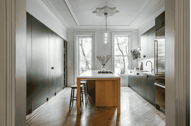 A refined galley-style kitchen viewed from an adjoining doorway, with full-height dark grey cabinetry on both sides, a central timber island with a white benchtop and two stools, integrated stainless-steel appliances, and a sink beneath tall windows. The room features polished herringbone timber flooring, white walls, ornate ceiling cornices and mouldings, a decorative ceiling medallion, and pendant lighting. The overall composition blends contemporary kitchen joinery with traditional architectural detailing.